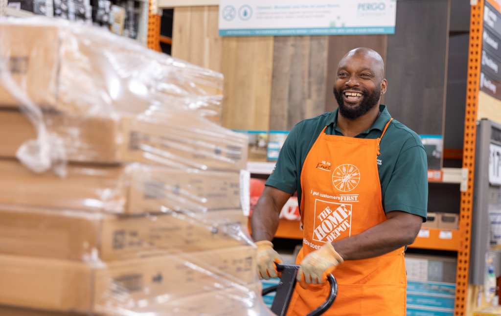 Middle-aged black man in a green polo and orange apron smiles as he transports boxes in a store.