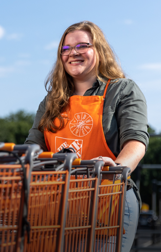 Woman in an orange Home Depot apron, smiling and pushing multiple Home Depot shopping carts